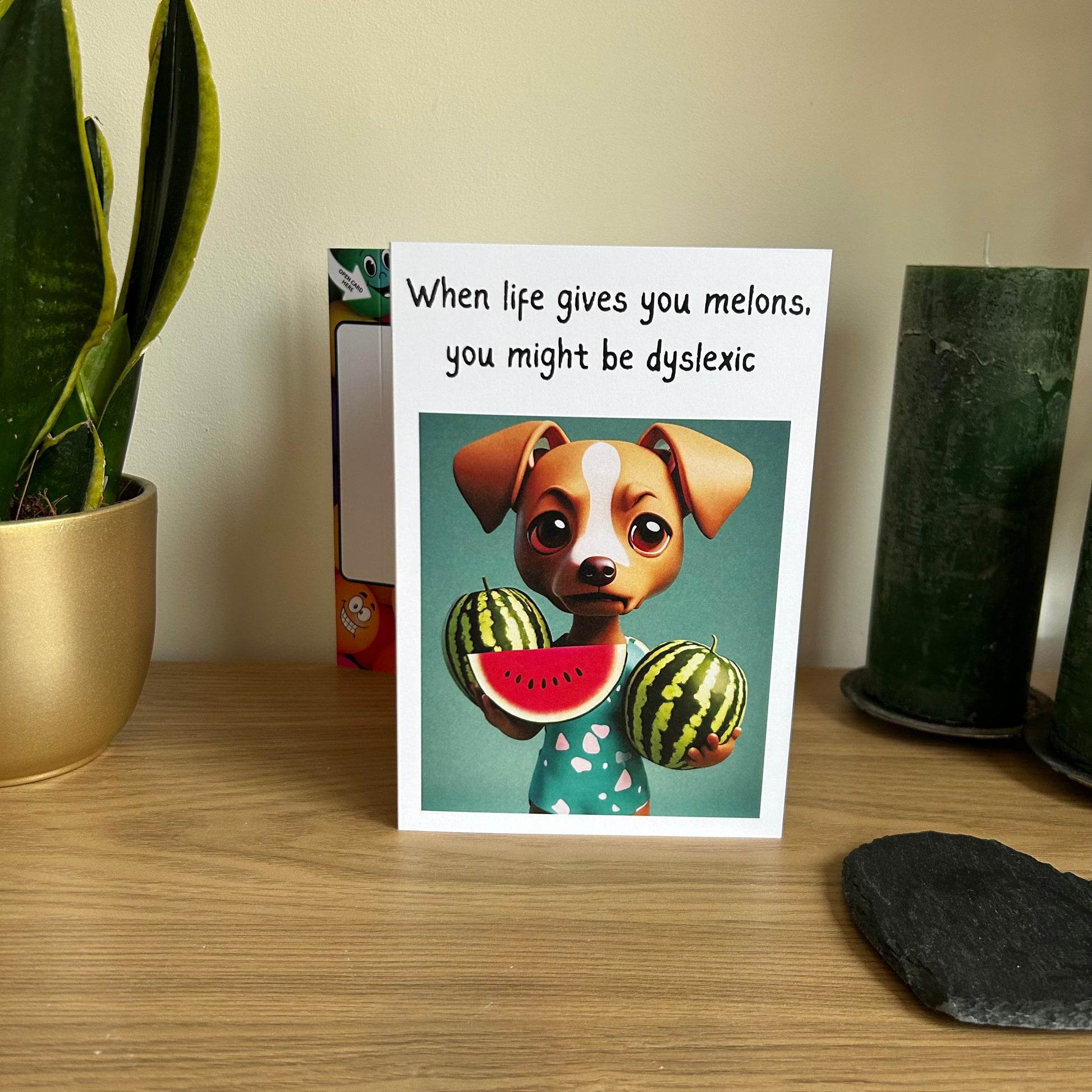Image shows the Bare Cards Giggle Balls Humour Range 'When Life Gives you Melons' birthday card displayed in a home setting. The illustration shows a cute cartoon dog holding 3 melons and the caption reads 'When life gives you melons, you might be dyslexic'