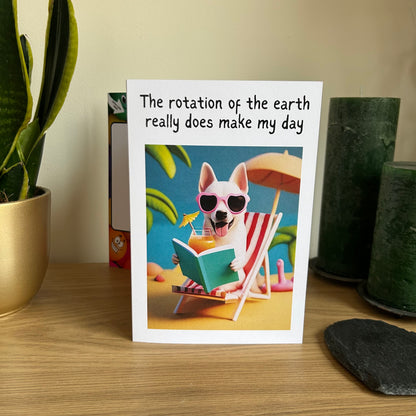 Image shows the Bare Cards Giggle Balls Humour Range 'Make my Day' Birthday Card displayed in a home setting. The illustration shows a cute cartoon dog sitting on a deckchair, wearing pink sunglasses and holding a cocktail and a book. The caption reads 'The rotation of the earth really does make my day'