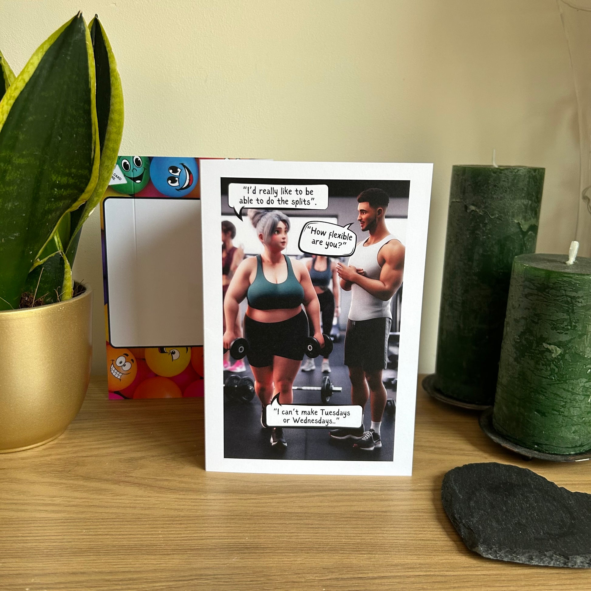 Image shows the Bare Cards Giggle Balls Humour Range 'Gym Joke' Birthday Card displayed in a home setting. The illustration shows a larger lady in gym gear holding dumbells taking to a fit gym instructor. She is saying "I'd really like to be able to do the splits". He replies "How flexible are you?" She replies "I can't make Tuesdays or Wednesdays.."
