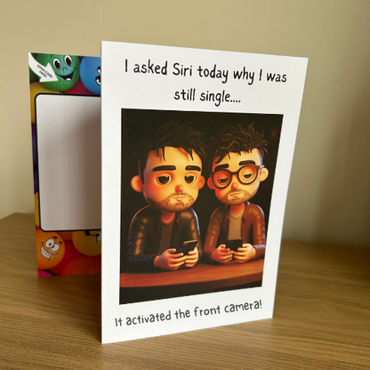 Image shows the Bare Cards Giggle Balls Humour Range 'Siri Joke' displayed in a home setting. The illustration shows two men looking glum and sitting down using their phones. The caption reads 'Iasked Siri today why I was still single.... It activated the front camera!'