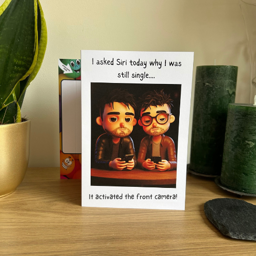 Image shows the Bare Cards Giggle Balls Humour Range 'Siri Joke' displayed in a home setting. The illustration shows two men looking glum and sitting down using their phones. The caption reads 'Iasked Siri today why I was still single.... It activated the front camera!'