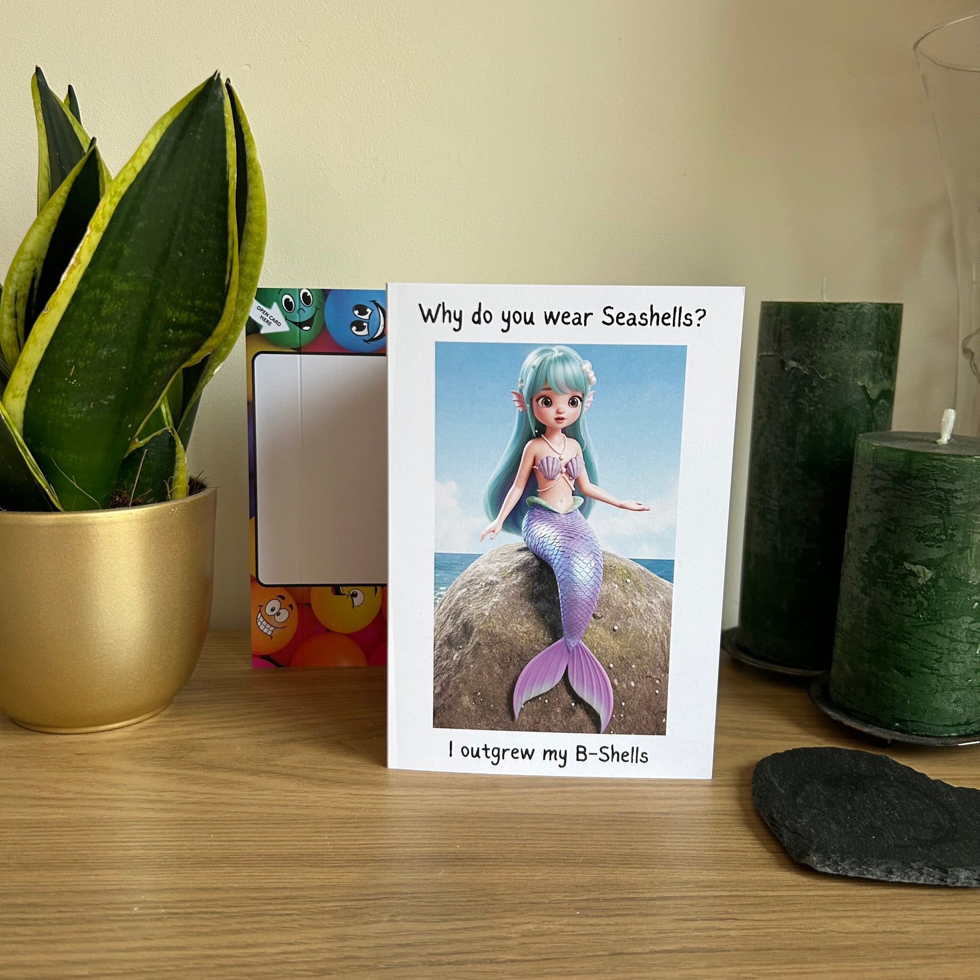 Image shows the Bare Cards Giggle Balls Humour Range 'Mermaid Birthday card' displayed in a home setting. The illustration shows a pretty cartoon mermaid with pastel blue hair and a lilac tail. She has lilac seashells covering her upper body for modesty. The top caption reads 'Why do you wear Seashells?'. The bottom caption reads 'I outgrew my B-Shells'