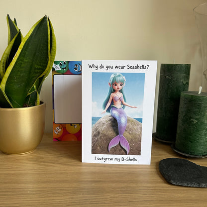 Image shows the Bare Cards Giggle Balls Humour Range 'Mermaid Birthday card' displayed in a home setting. The illustration shows a pretty cartoon mermaid with pastel blue hair and a lilac tail. She has lilac seashells covering her upper body for modesty. The top caption reads 'Why do you wear Seashells?'. The bottom caption reads 'I outgrew my B-Shells'