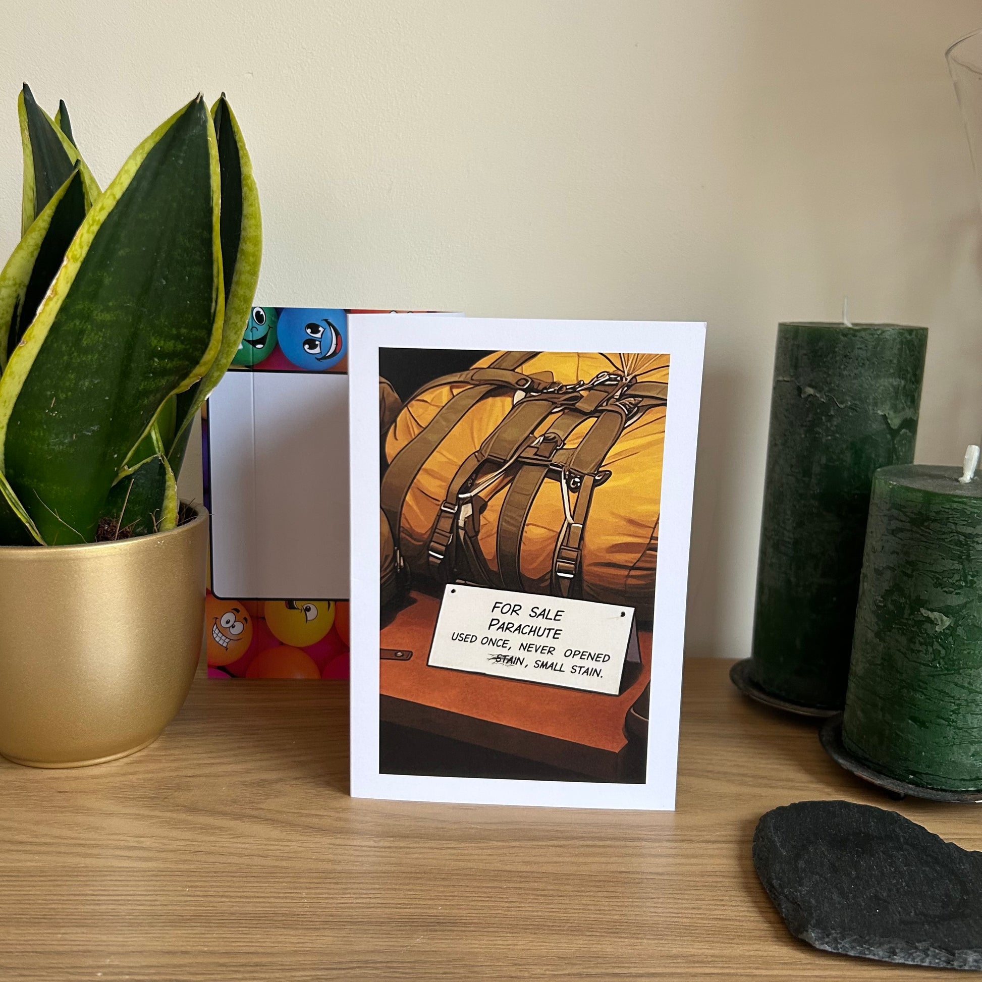 Image show the Bare Cards Giggle Balls Humour Range 'Parachute For Sale' Birthday card displayed in a home ssetting. The illustration shows a packed up yellow parachute in front of a for sale sign which reads 'FOR SALE PARACHUTE, USED ONCE, NEVER OPENED, STAIN, SMALL STAIN'  The first word stain is scribbled out.