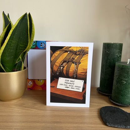 Image show the Bare Cards Giggle Balls Humour Range 'Parachute For Sale' Birthday card displayed in a home ssetting. The illustration shows a packed up yellow parachute in front of a for sale sign which reads 'FOR SALE PARACHUTE, USED ONCE, NEVER OPENED, STAIN, SMALL STAIN'  The first word stain is scribbled out.