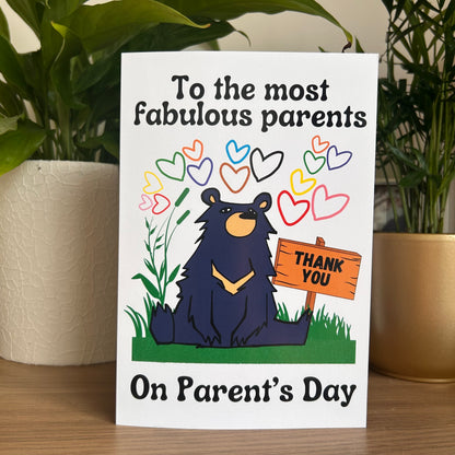 The front of a white greeting card with a cartoon black bear sitting on grass and holding a wooden sign that says THANK YOU. The card reads 'To the most fabulous parents On Parent's Day' and is photographed standing on a wooden surface between two potted green plants.