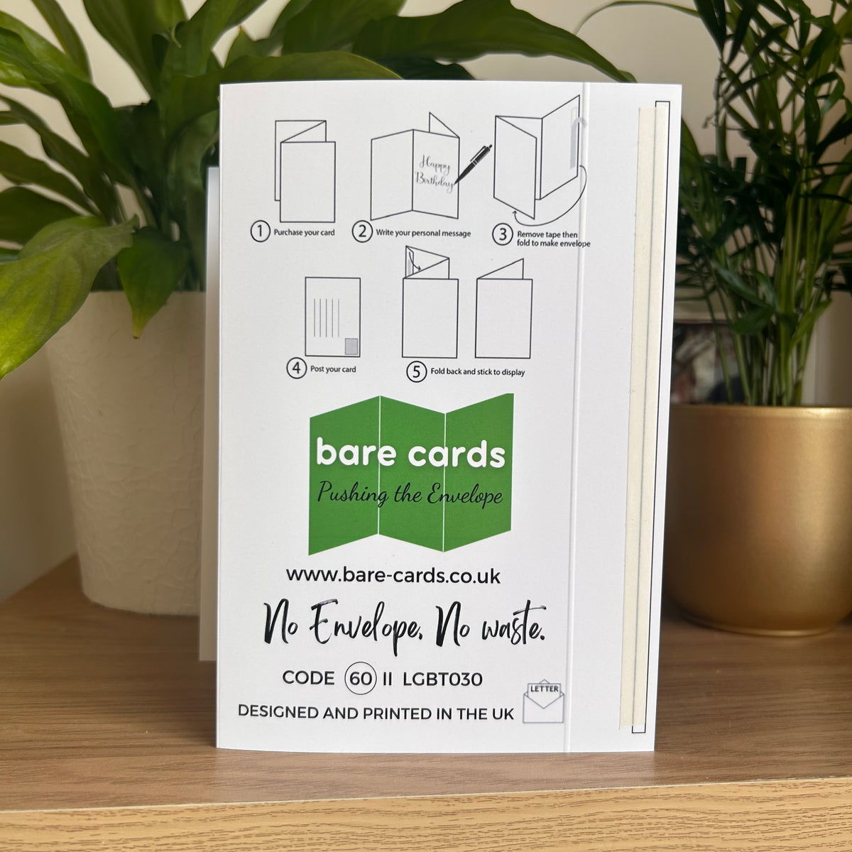 The back of the Bare Cards Happy Parent's Day self-sealing card showing step-by-step envelope folding instructions, photographed standing on a wooden surface between two potted green plants.