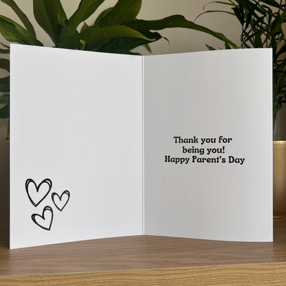 An open white greeting card on a wooden surface, with three black outlined hearts on the left panel and the text 'Thank you for being you! Happy Parent's Day' on the right panel. Green plants and a gold vase are visible in the background.