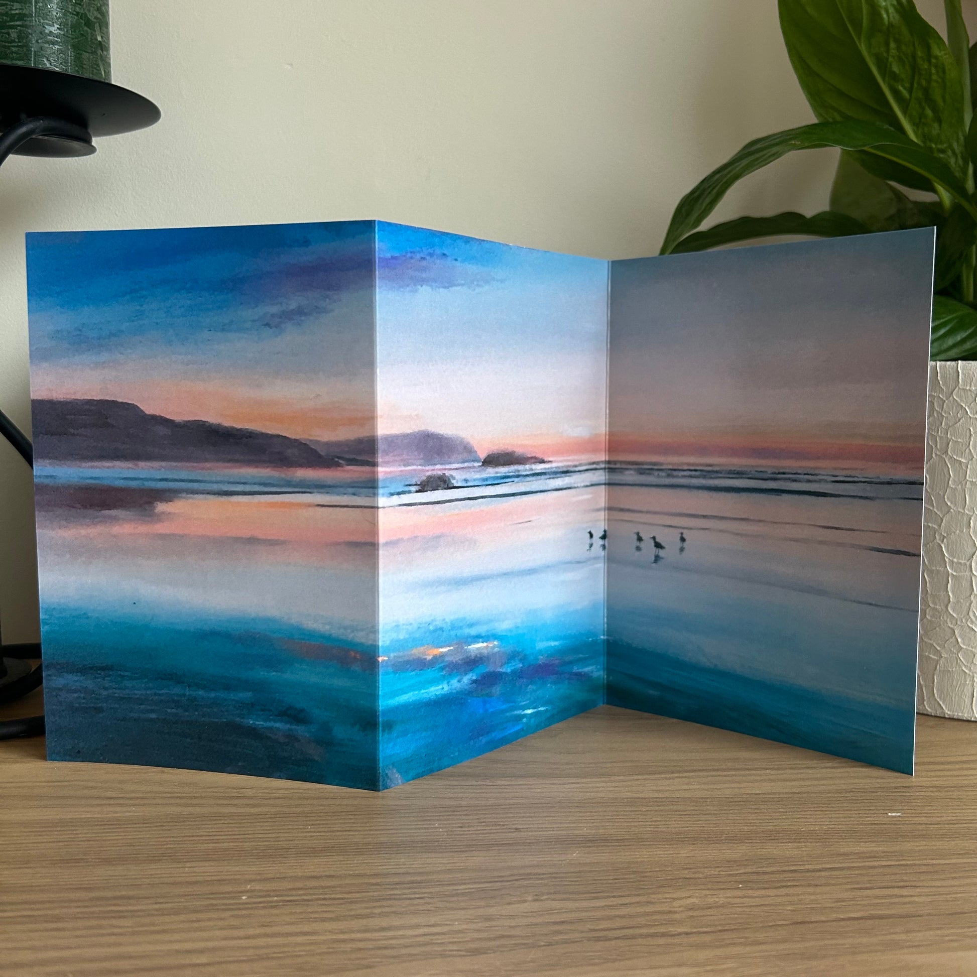 Folded trifold card with a scenic illustration of a sunset over Polzeath beach on a wooden surface.