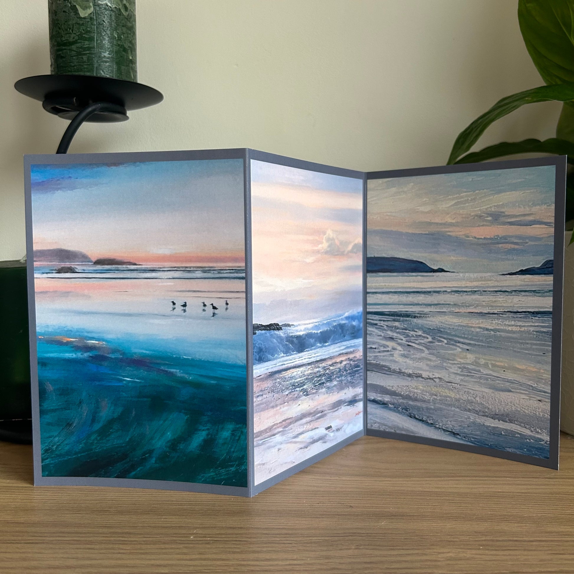 Foldable trifold greetings card with a triptych of paintings showing scenic designs of a beach and ocean on a wooden surface.