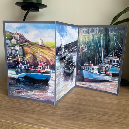 Tri-fold card with triptych paintings of boats and coastal scenes on a wooden surface.