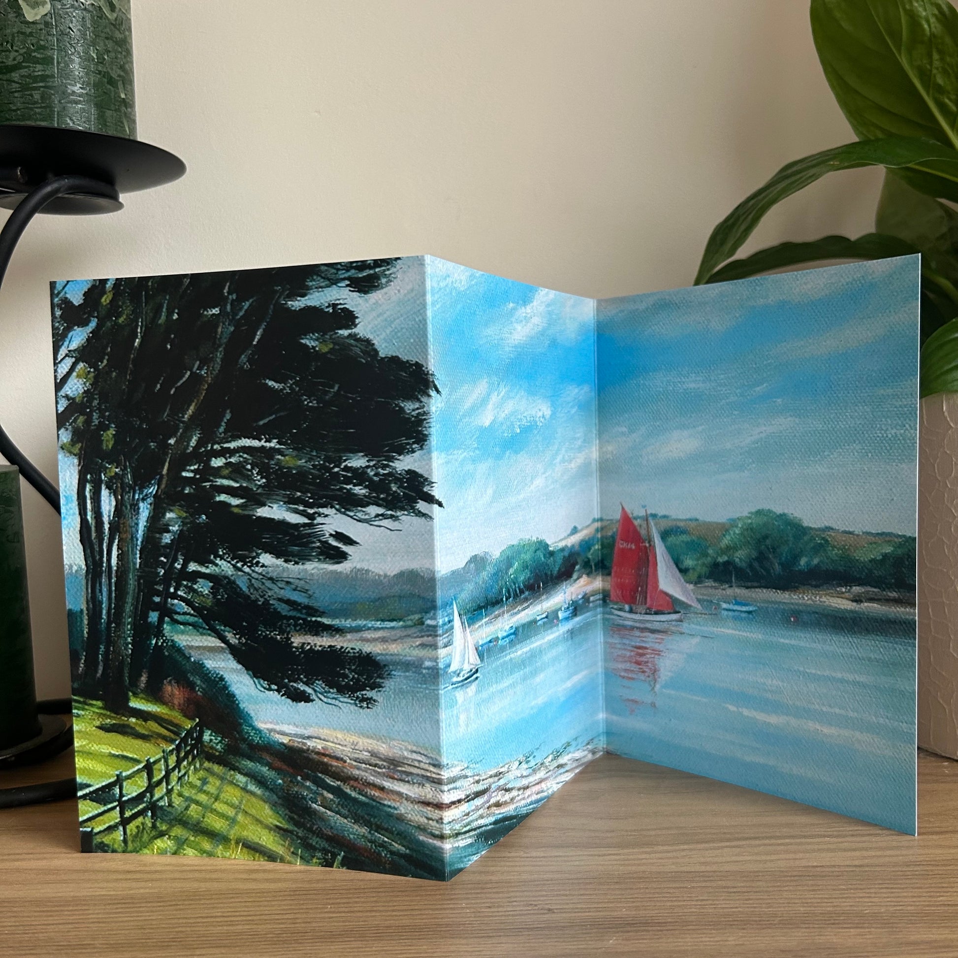 Tri-fold card with a scenic painting of a river and sailboats on a wooden surface.