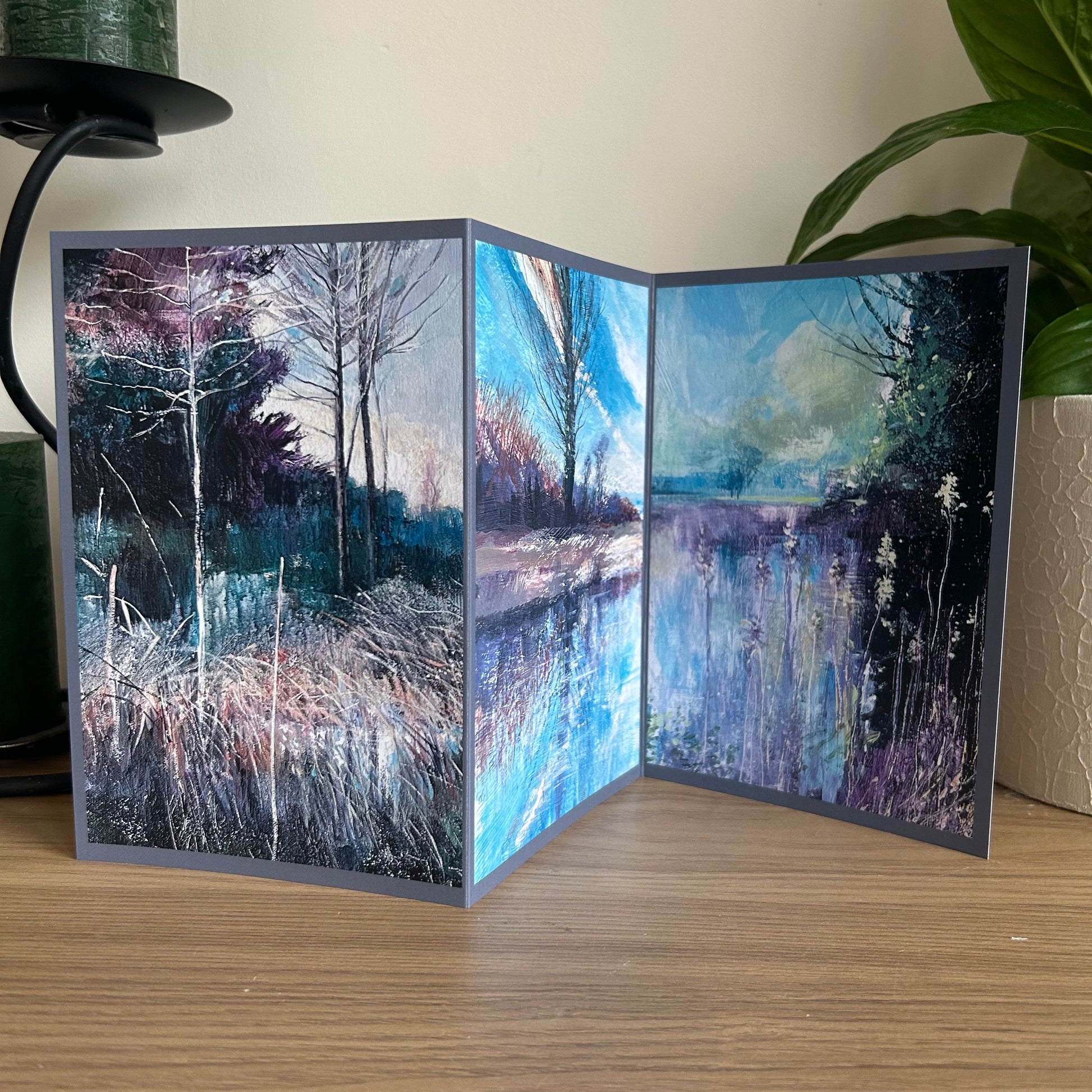 Greetings card showing a triptych landscape painting on a wooden surface with a plant in the background