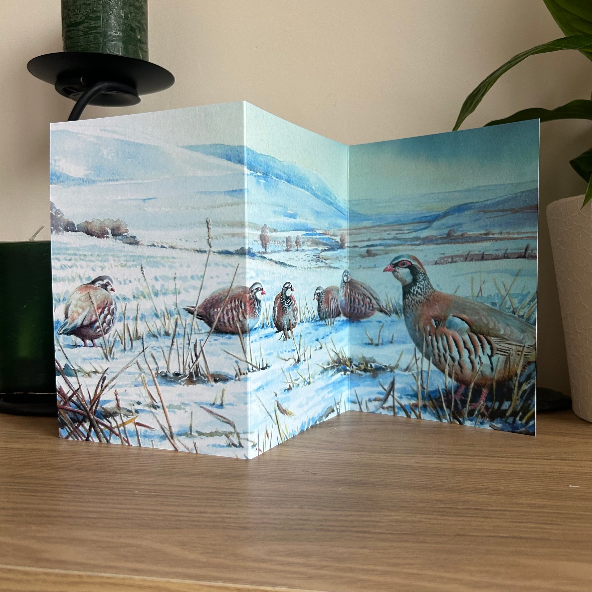 Trifold Greetings Card with a winter landscape and partridge illustration on a wooden surface.