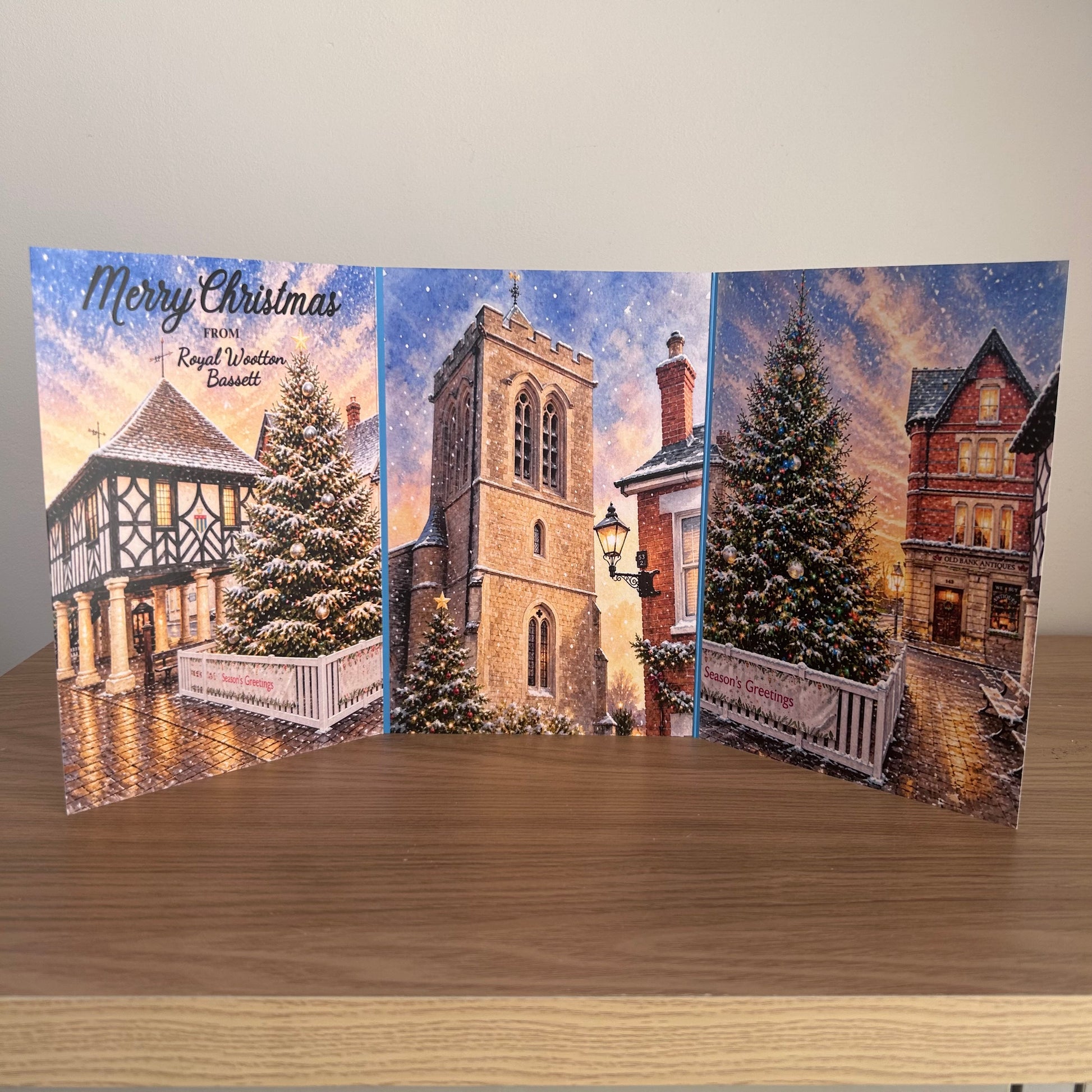 A three-panel Christmas greeting card displayed open on a wooden surface against a plain light wall. The card features winter village scenes with snow-covered trees, historic buildings, and the text 'Merry Christmas from Royal Wootton Bassett'.