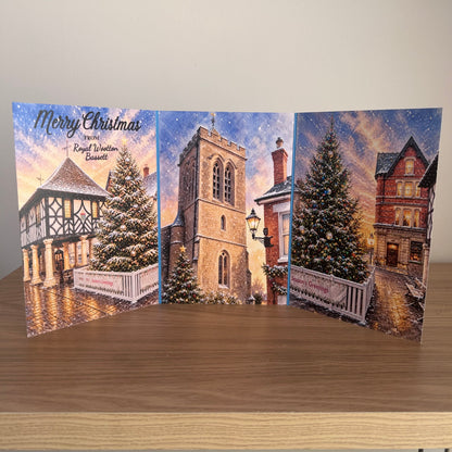 A three-panel Christmas greeting card displayed open on a wooden surface against a plain light wall. The card features winter village scenes with snow-covered trees, historic buildings, and the text 'Merry Christmas from Royal Wootton Bassett'.