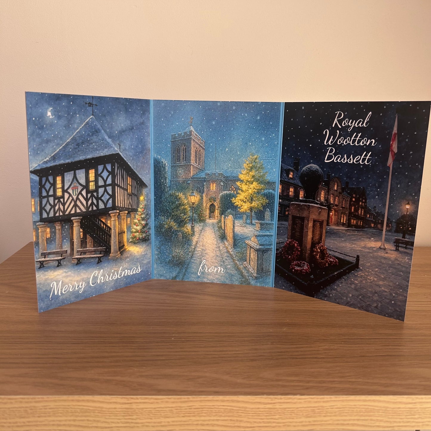 A three-panel Christmas greeting card set displayed on a wooden surface against a beige wall. The left panel shows a snowy town hall with the text 'Merry Christmas'; the center panel depicts a church street at night with snow falling and the word 'from'; the right panel shows a snowy town square with a monument and the text 'Royal Wootton Bassett'.