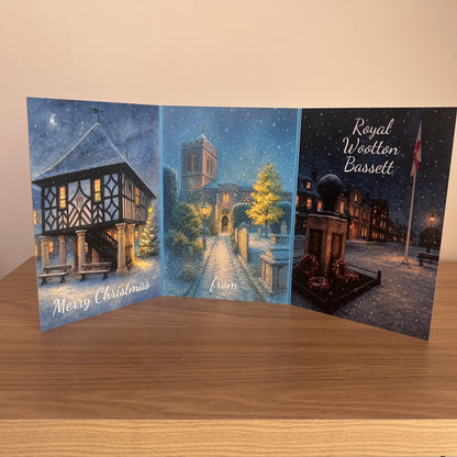 A three-panel Christmas greeting card set displayed on a wooden surface against a beige wall. The left panel shows a snowy town hall with the text 'Merry Christmas'; the center panel depicts a church street at night with snow falling and the word 'from'; the right panel shows a snowy town square with a monument and the text 'Royal Wootton Bassett'.