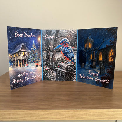 A triptych Christmas greeting card displayed standing upright on a wooden table against a beige wall. The left panel shows a snowy house and Christmas tree with the text 'Best Wishes and a very Merry Christmas'; the center panel shows a blue and red kingfisher in a snowy outdoor scene with 'from'; the right panel shows a church at night with 'Royal Wootton Bassett'.