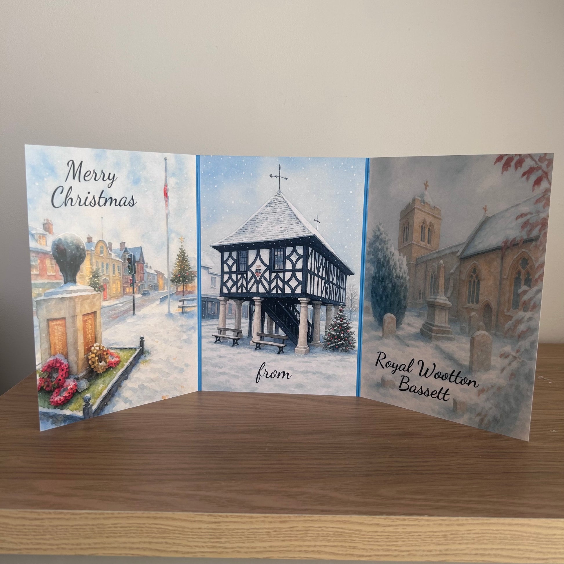 A triptych Christmas card displayed on a wooden table against a plain wall. The card features three illustrated panels showing a snowy street scene, a timber-frame structure, and a churchyard, with text reading 'Merry Christmas', 'from', and 'Royal Wootton Bassett'.