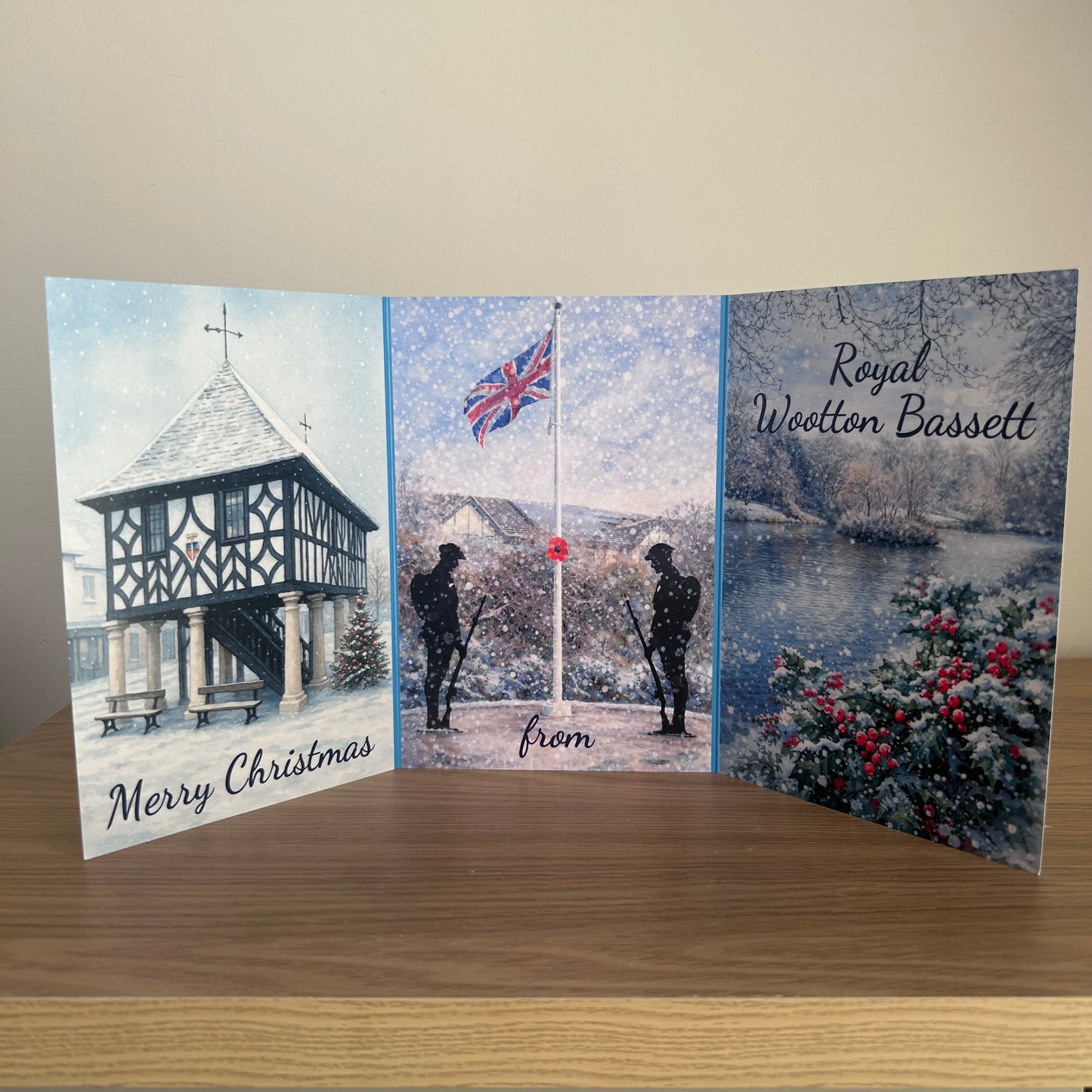 A  Christmas memorial tri-fold card displayed on a wooden table against a beige wall. The card features three illustrated panels: the left panel shows a winter scene of a timber-framed gazebo with 'Merry Christmas' written at the bottom; the center panel shows soldiers standing at a flagpole in snow with 'from' written below; the right panel shows a snowy landscape with a lake and 'Royal Wootton Bassett' written at the top.