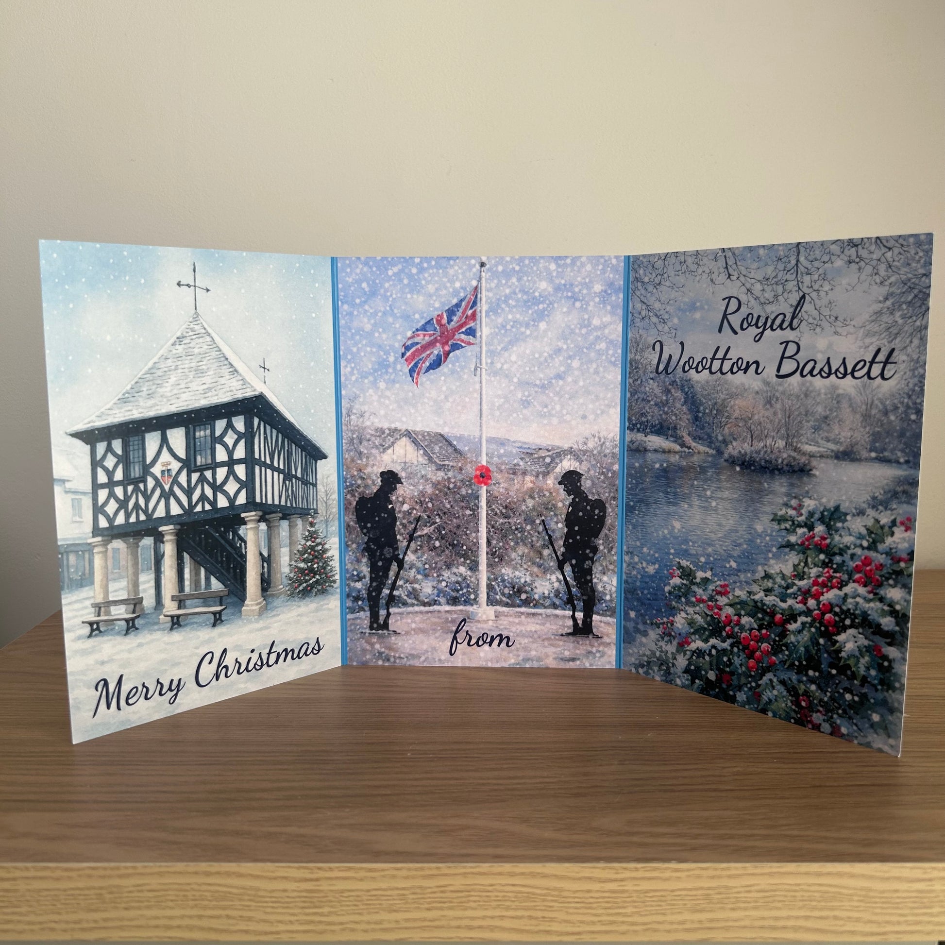 A  Christmas memorial tri-fold card displayed on a wooden table against a beige wall. The card features three illustrated panels: the left panel shows a winter scene of a timber-framed gazebo with 'Merry Christmas' written at the bottom; the center panel shows soldiers standing at a flagpole in snow with 'from' written below; the right panel shows a snowy landscape with a lake and 'Royal Wootton Bassett' written at the top.
