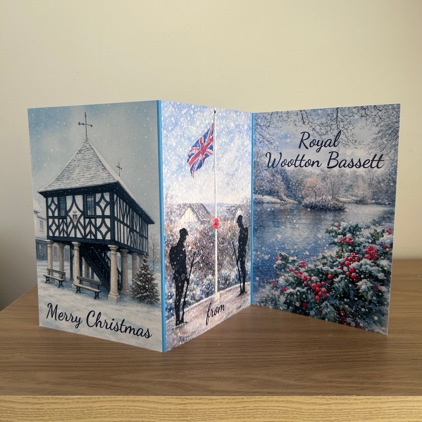 A triptych folding Christmas greeting card displayed on a wooden table against a beige wall. The card features three panels with winter scenes: a snow-covered town hall on the left, silhouettes under a Union Jack flag in the middle, and a snowy river landscape with red berries on the right.