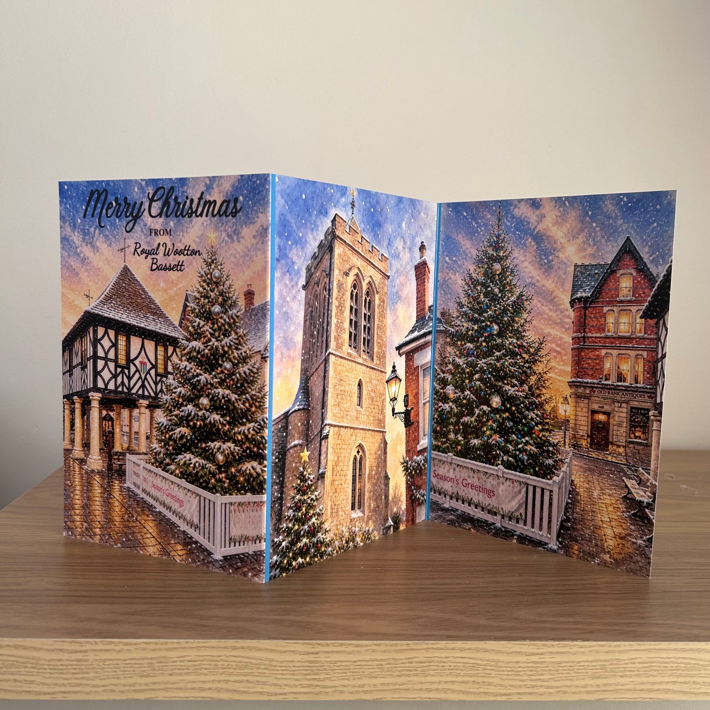 A Triptych Christmas greeting card displayed on a wooden table against a plain beige wall. The card features a winter village scene with snow-covered Christmas trees, historic houses, and a church tower under a starry night sky with the text 'Merry Christmas from Royal Wootton Bassett'.