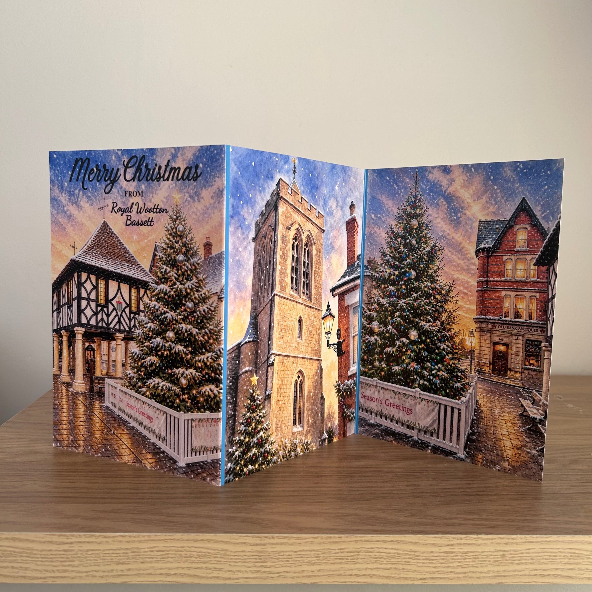 A Triptych Christmas greeting card displayed on a wooden table against a plain beige wall. The card features a winter village scene with snow-covered Christmas trees, historic houses, and a church tower under a starry night sky with the text 'Merry Christmas from Royal Wootton Bassett'.