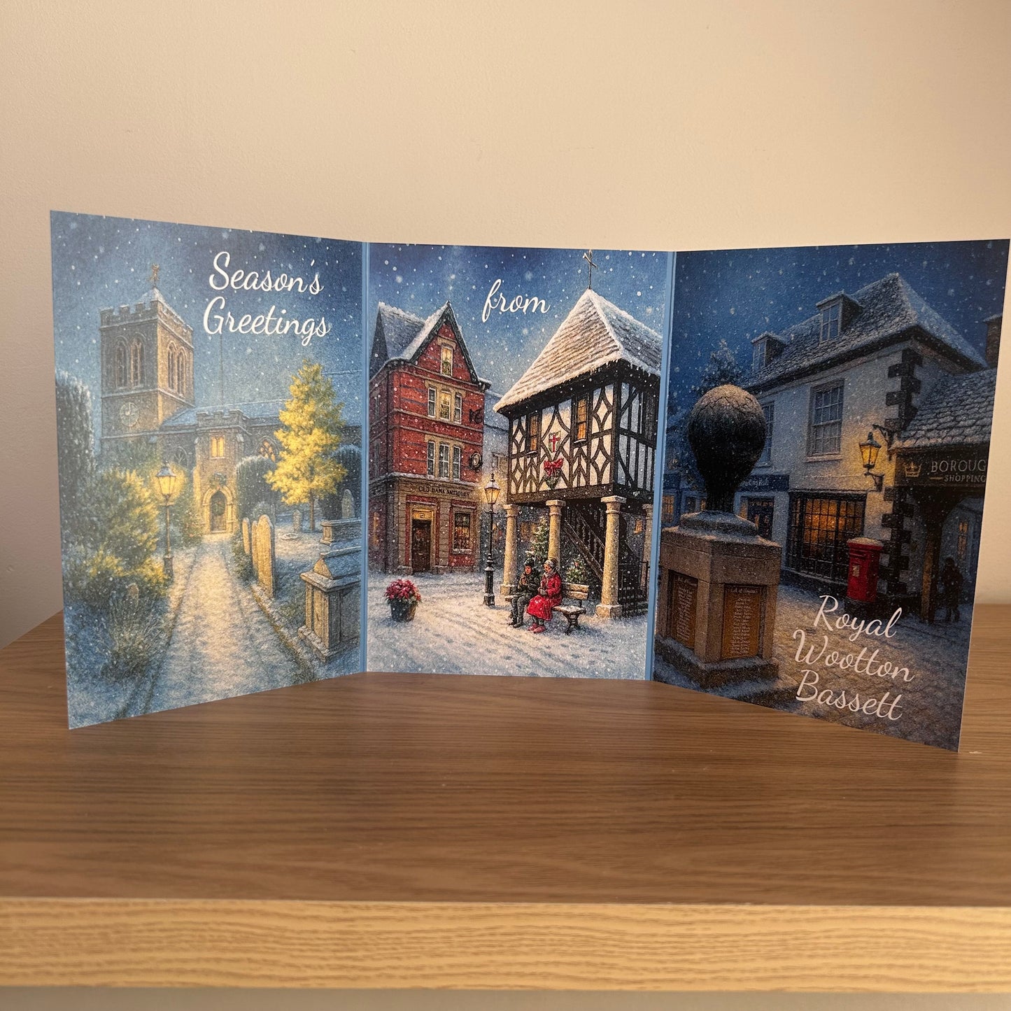 A three-panel Christmas greeting card standing open on a wooden surface against a beige wall. The card depicts a snowy winter street scene with buildings, lampposts, and the text 'Season's Greetings' and 'from Royal Wootton Bassett'.