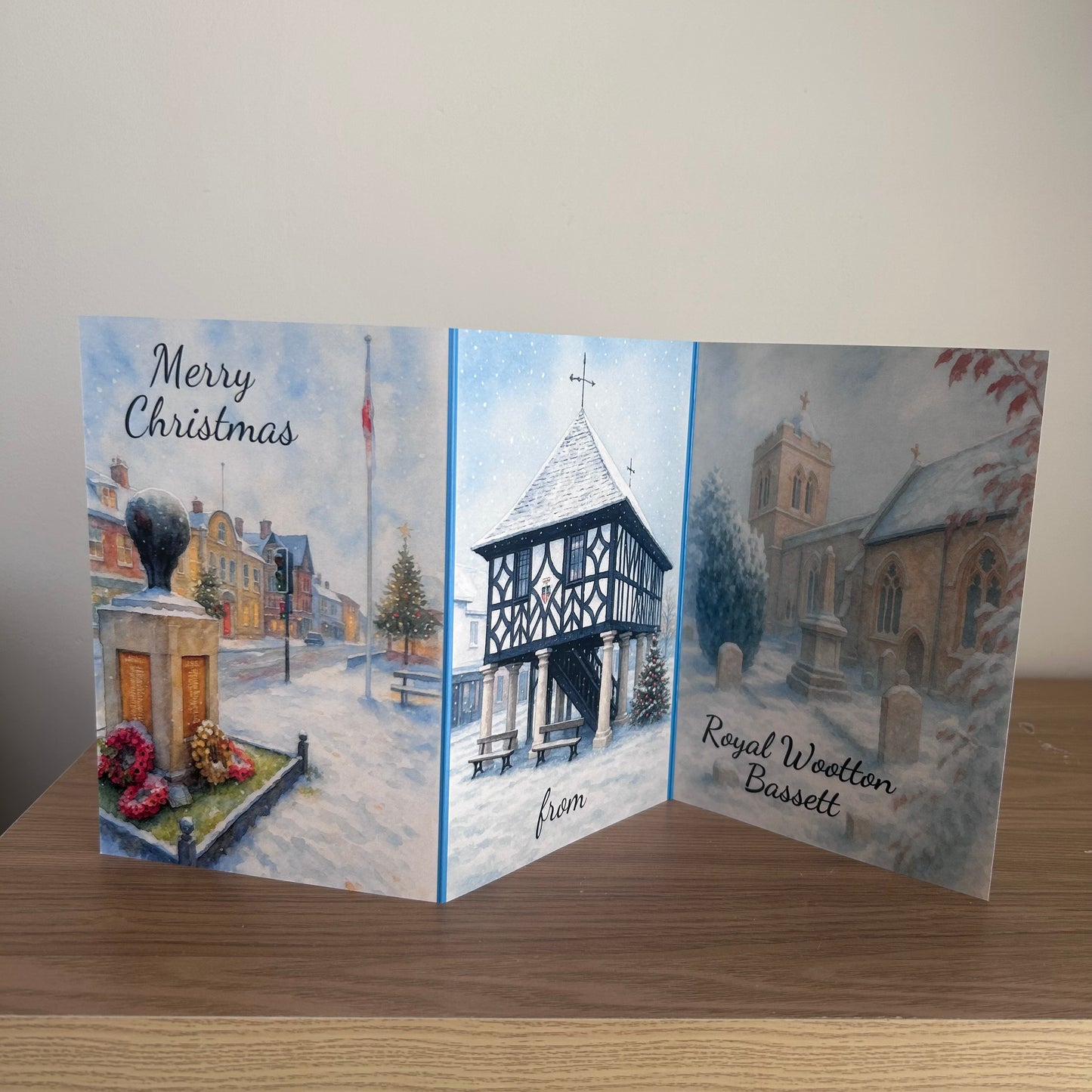 A three-panel folding Christmas greeting card displayed on a wooden surface against a plain light wall. The card features watercolor winter scenes: the left panel shows a snowy street with a monument and the words 'Merry Christmas'; the center panel depicts a snow-covered timber-frame building; the right panel shows a church in winter with the text 'Royal Wootton Bassett'.