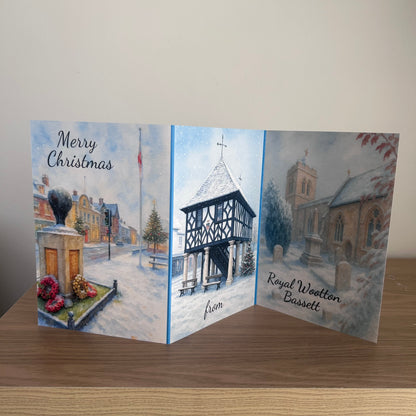 A three-panel folding Christmas greeting card displayed on a wooden surface against a plain light wall. The card features watercolor winter scenes: the left panel shows a snowy street with a monument and the words 'Merry Christmas'; the center panel depicts a snow-covered timber-frame building; the right panel shows a church in winter with the text 'Royal Wootton Bassett'.