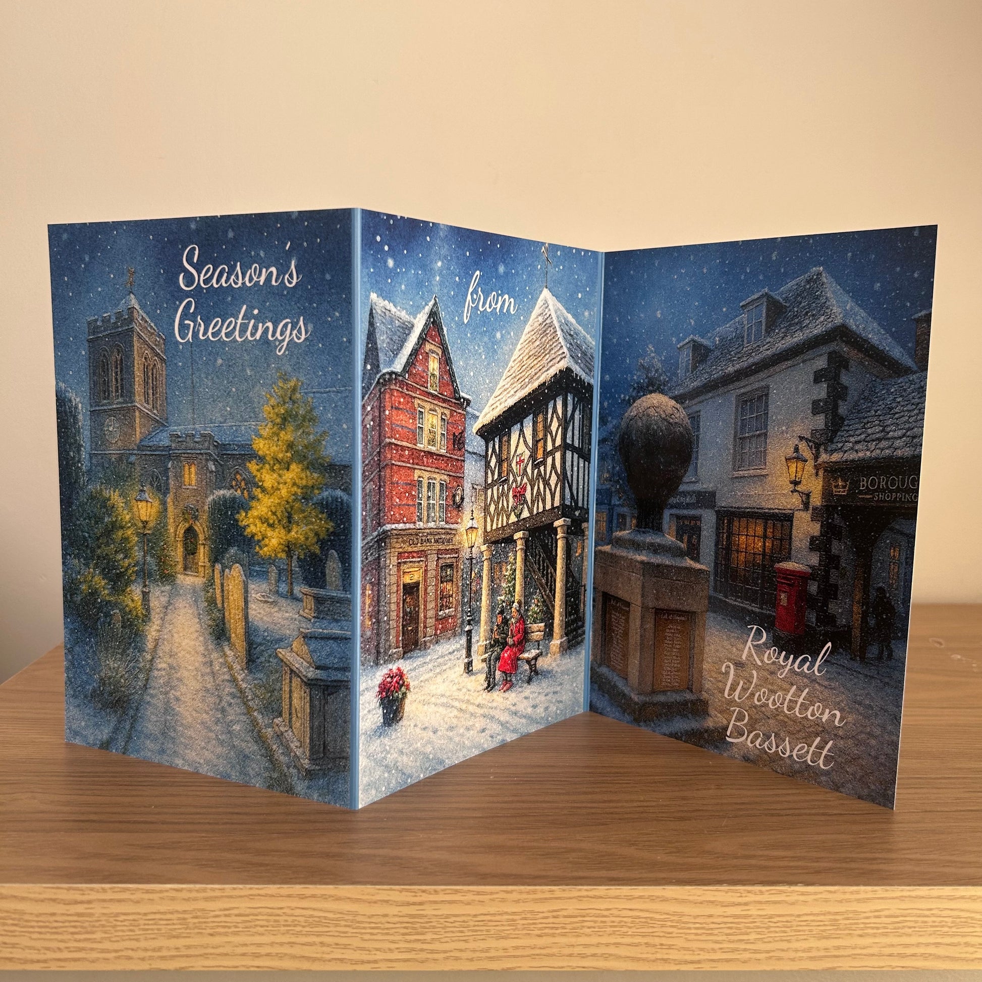 A three-panel accordion-style greeting card displayed open on a wooden surface against a beige wall. The card features a snowy village scene with text reading 'Season's Greetings', 'from', and 'Royal Wootton Bassett'.