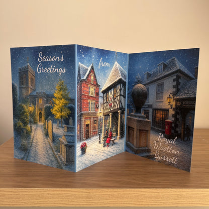 A three-panel accordion-style greeting card displayed open on a wooden surface against a beige wall. The card features a snowy village scene with text reading 'Season's Greetings', 'from', and 'Royal Wootton Bassett'.