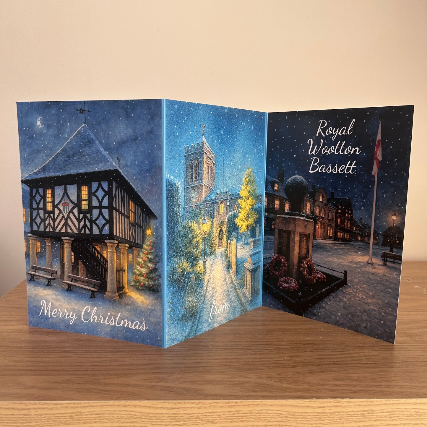 A three-panel folding Christmas card displayed open on a wooden surface against a beige wall. The card features winter town scenes: a town hall on pillars, a church street scene in the center, and a monument with the text 'Merry Christmas from Royal Wootton Bassett'