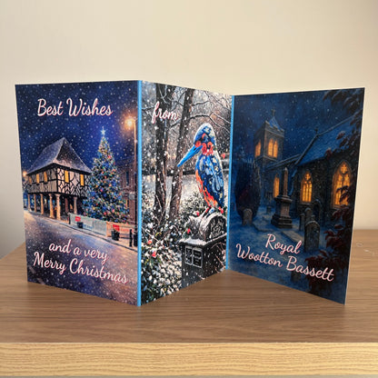 A handmade folding Christmas greeting card displayed open on a wooden table against a beige wall. The card features three panels with winter village scenes: Town Hall and Christmas tree on the left, a kingfisher on a postman box in the middle, and a snowy church with glowing windows on the right. Text reads: Best Wishes and a very Merry Christmas from Royal Wootton Bassett