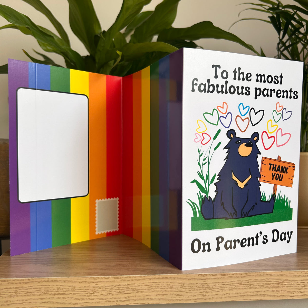 An open self-sealing parent's day greeting card displayed on a wooden surface with a green plant in the background. The left panel features a rainbow stripe design with a blank white address box, and the right panel is white with the text 'To the most fabulous parents On Parent's Day' and an illustration of a black bear holding a 'THANK YOU' sign.