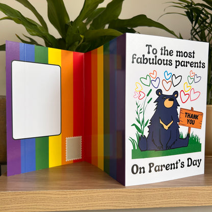 An open self-sealing parent's day greeting card displayed on a wooden surface with a green plant in the background. The left panel features a rainbow stripe design with a blank white address box, and the right panel is white with the text 'To the most fabulous parents On Parent's Day' and an illustration of a black bear holding a 'THANK YOU' sign.