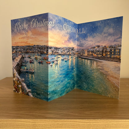 A Christmas greeting card featuring a scenic watercolor painting of a harbor in Cornwall, with boats, buildings, and a starry night sky. The card is standing open on a wooden surface against a beige wall, with 'Merry Christmas from CORNWALL' printed on the front.