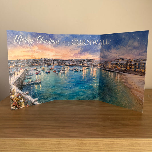 A Trifold Christmas greeting card featuring a scenic watercolor painting of a harbor in Cornwall under a snowy evening sky, displayed on a wooden surface against a beige wall.