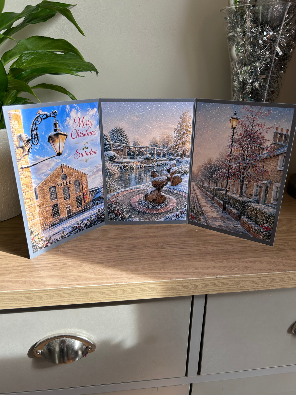 A three-panel Swindon Christmas greeting card displayed on a wooden cabinet, featuring festive images of the Steam Museum, Queen's Park and The Railway Village. The left panel includes the text 'Merry Christmas from Swindon'.