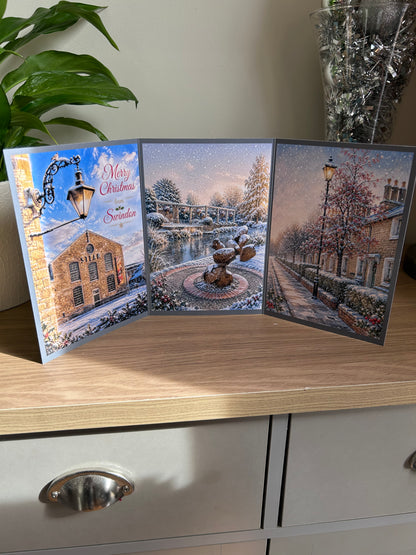 A three-panel Swindon Christmas greeting card displayed on a wooden cabinet, featuring festive images of the Steam Museum, Queen's Park and The Railway Village. The left panel includes the text 'Merry Christmas from Swindon'.