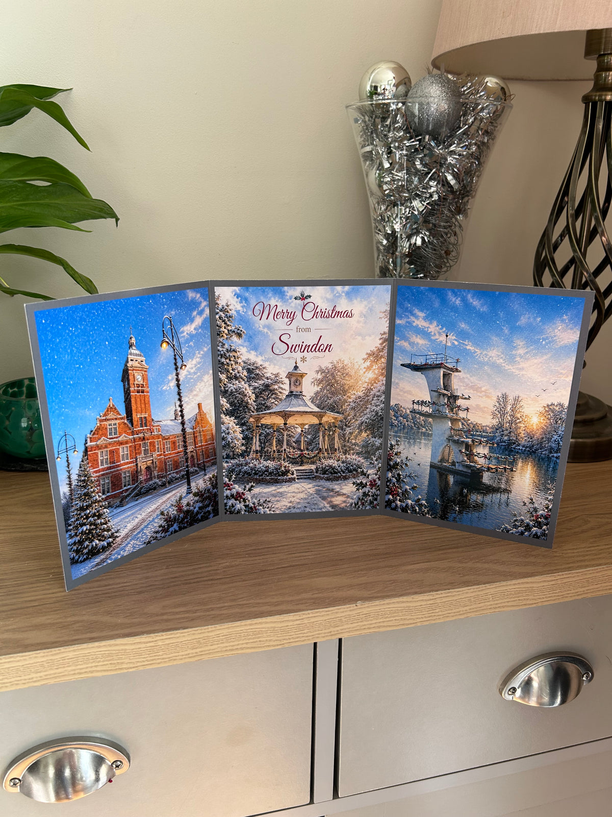 A three-panel Christmas greeting card displayed on a wooden cabinet, featuring winter scenes: the Town Hall, a snowy garden pavilion in Town Gardens with the text 'Merry Christmas from Swindon,' and the diving board at Coate Water at sunset. Decorative items and a lamp are visible in the background.
