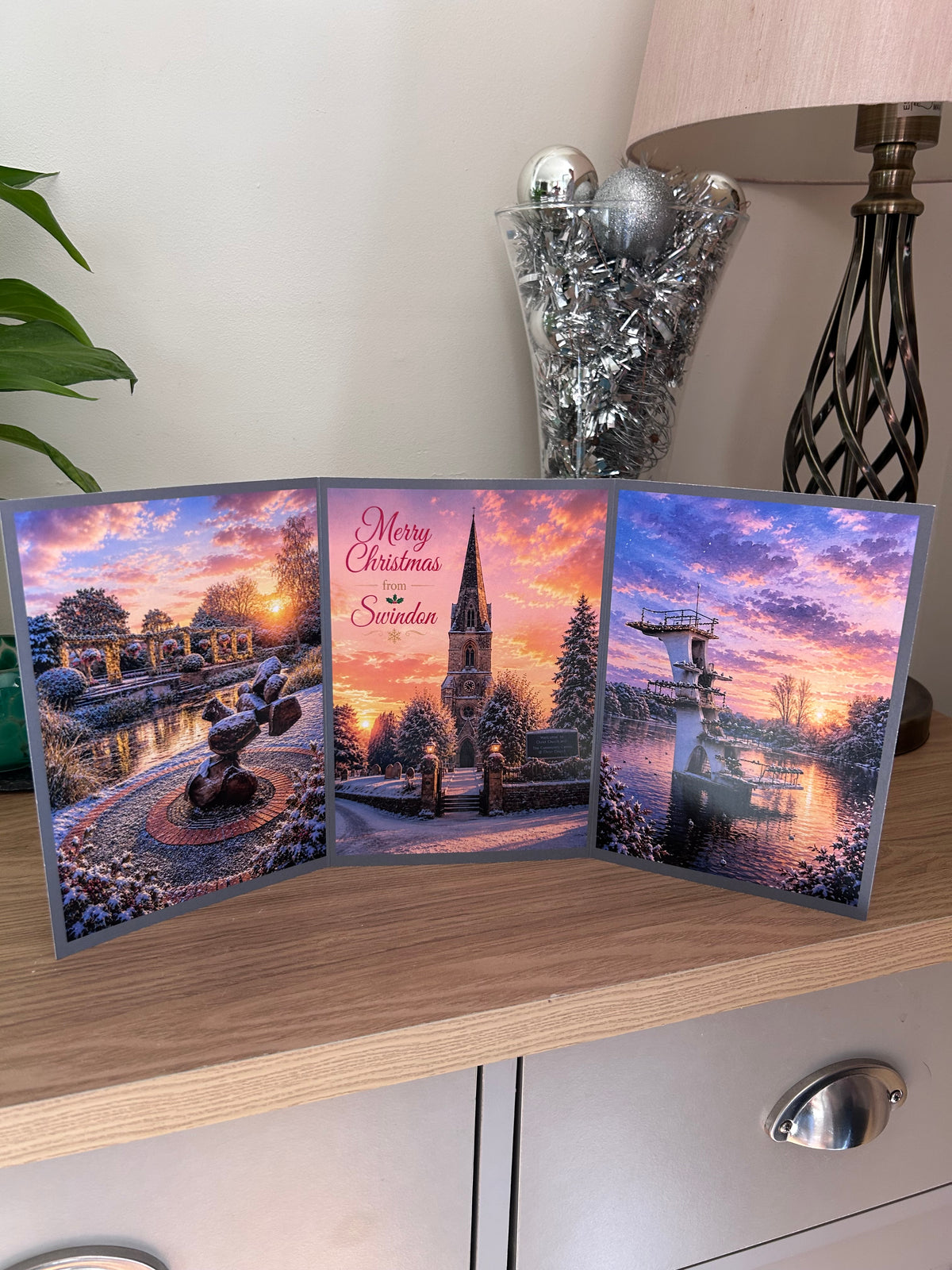A three-panel Swindon Christmas greeting card set displayed on a wooden cabinet, featuring scenic winter landscapes from Old Town Gardens, Christchurch and the Coate Water diving board under a colorful sunset sky.