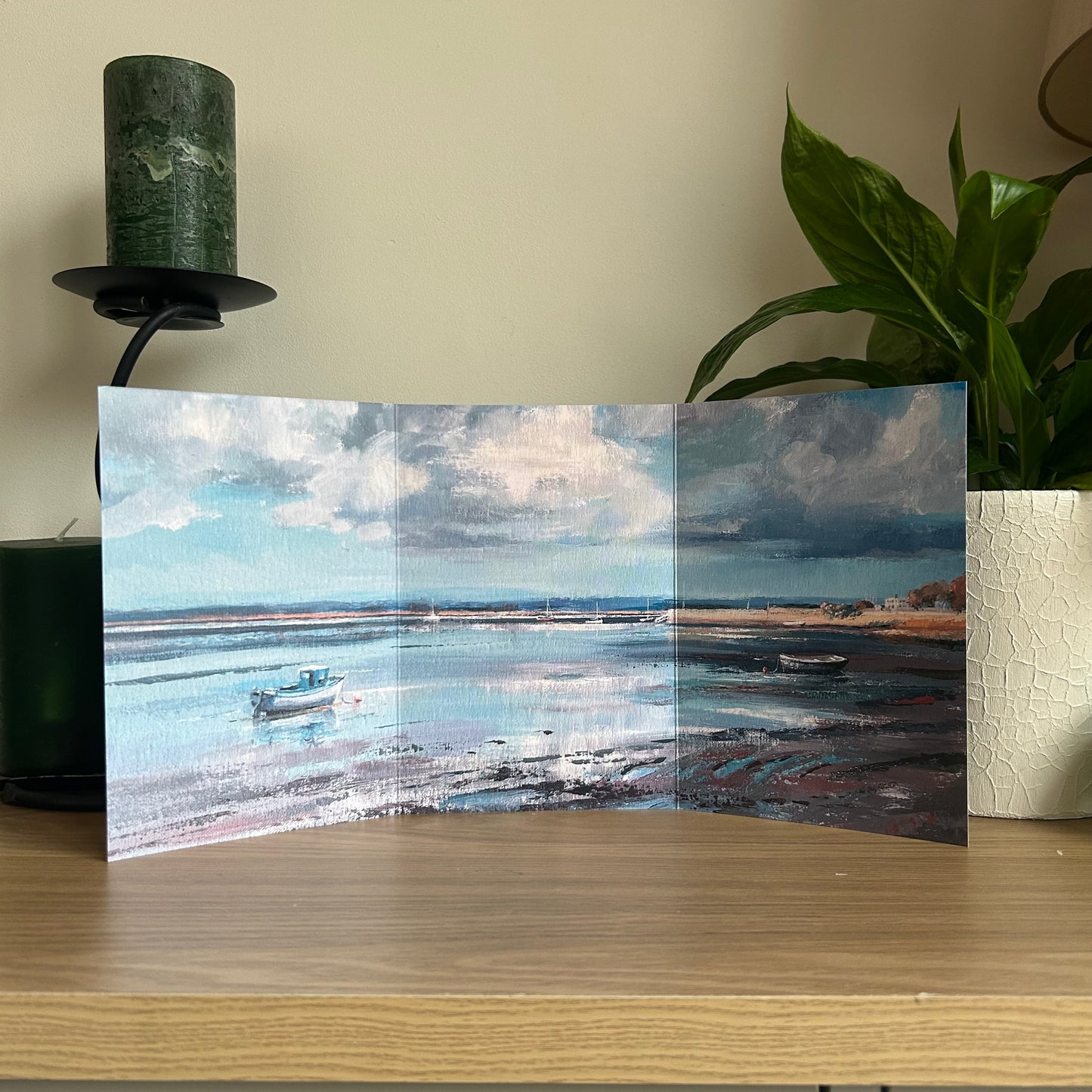 Self-sealing trifold greetings card with a scenic illustration of a beach and boats on a wooden surface with a plant and candle in the background.