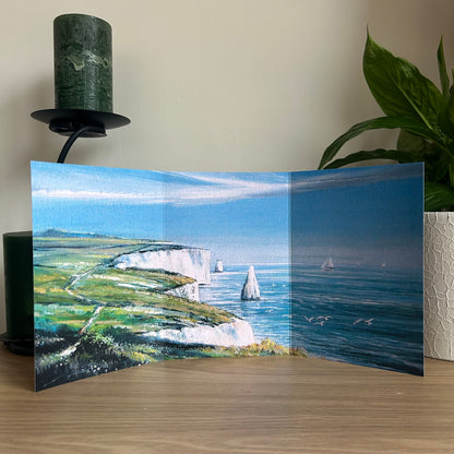 Foldable scenic card with a coastal landscape on a wooden surface with a plant and candle in the background.