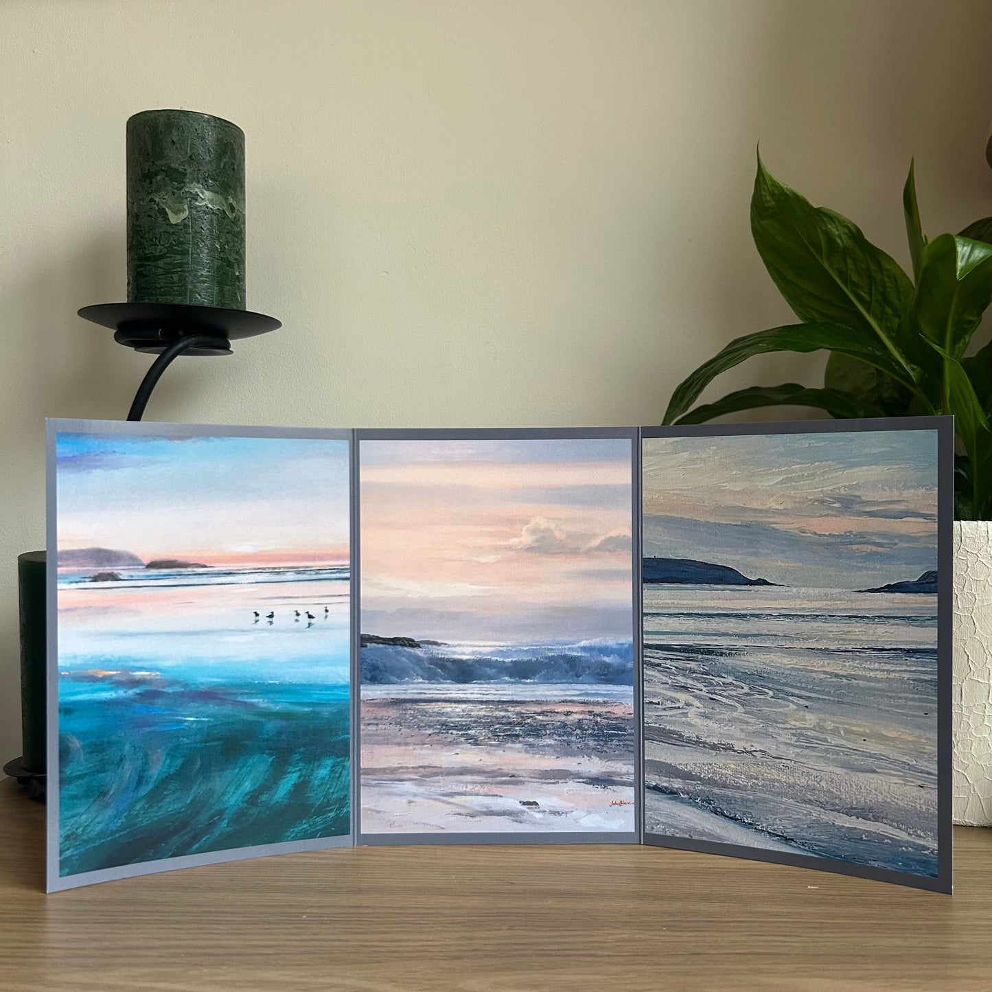 Triptych greetings card with three beach scene paintings on a wooden surface with a plant and candle in the background.
