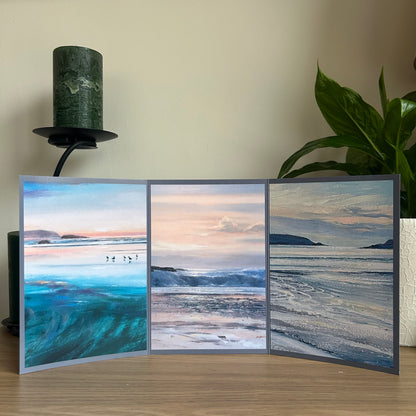 Triptych greetings card with three beach scene paintings on a wooden surface with a plant and candle in the background.