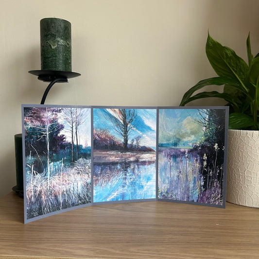Greetings card showing a triptych painting of nature scenes displayed on a wooden surface with a candle and plant in the background.
