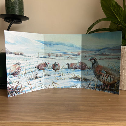 Trifold greetings card featuring a painting of partridges on the downs displayed on a wooden surface.